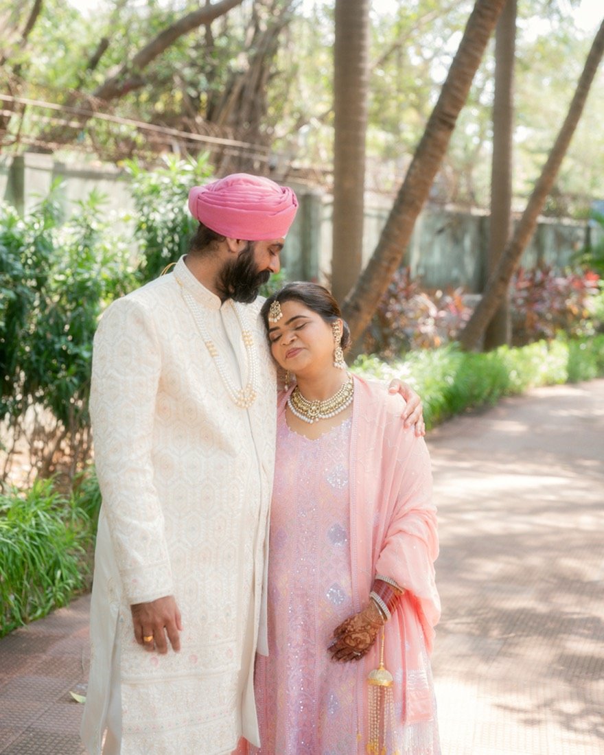 A groom in a pink turban holding the bride close beneath the trees
