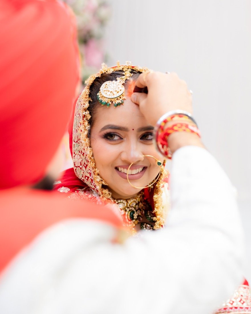 A bride adjusting her maang tikka, the groom's turban in the foreground
